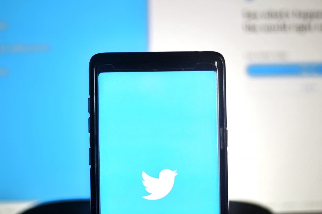 A photo of a smartphone displaying the Twitter logo in front of a blurred computer screen in the background.